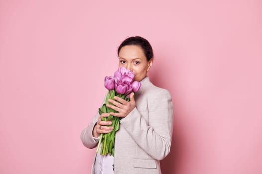 Attractive pregnant woman sniffing a beautiful bouquet of purple tulips, isolated over pink background. Mother's Day