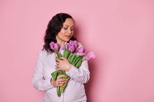 Delightful brunette, pregnant woman holding bunch of tulips on isolated pink background. Spring and femininity concept
