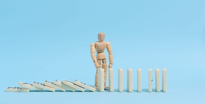 A wooden puppet toy holds back a falling domino on a blue background, representing the concept of a strong personality