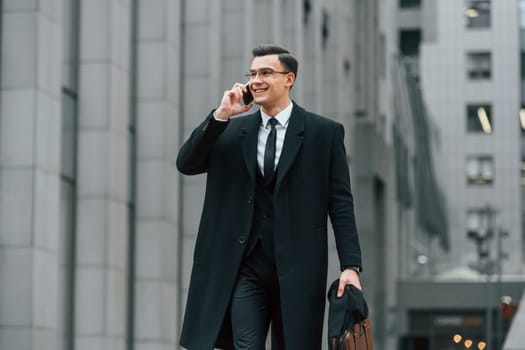 With smartphone. Businessman in black suit and tie is outdoors in the city