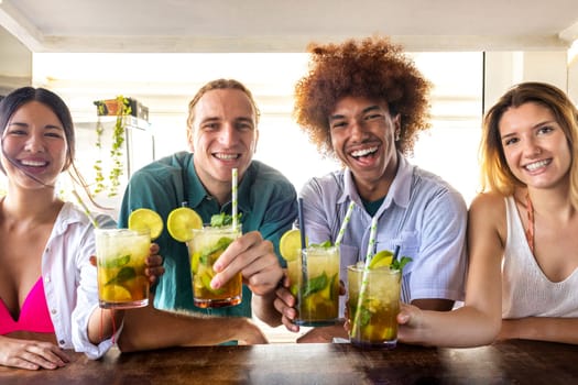Group of multiracial friends toasting with mojito cocktails at beach bar looking at camera. Happy friends having fun at summertime. Summertime lifestyle concept.