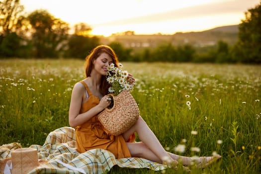 beautiful red-haired woman sits in a chamomile field on a plaid with a basket of flowers during sunset