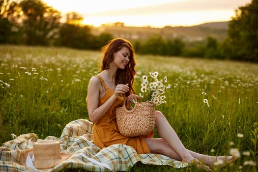 beautiful red-haired woman sits in a chamomile field on a plaid with a basket of flowers during sunset