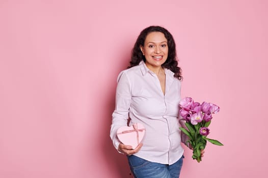 Attractive positive Latin American pregnant woman smiles looking at camera, posing with gift box and bouquet of tulips