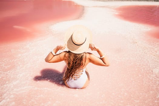 A woman traveler looks at an amazing pink salt lake. He sits with his back in a bathing suit and holds his hat in his hands. Wanderlust travel concept
