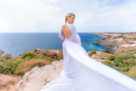 Blonde with long hair on a sunny seashore in a white flowing dress, rear view, silk fabric waving in the wind. Against the backdrop of the blue sky and mountains on the seashore.
