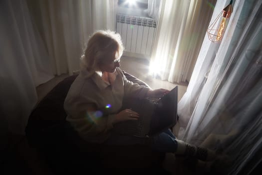 Silhouette and shadow of Adult mature woman of 40-60 years old with laptop, notebook, computer in warm sweater in dark calm cozy evening atmosphere room. Interior with curtains and small linght