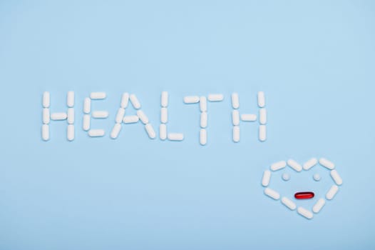 Word HEALTH and heart shape as emoticon laid out with medical therapeutic pills and translucent gel capsules, on isolated blue background. World Health Day. Pharmaceutical industry concept. Copy space