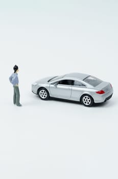 On a white surface is a car and a miniature figurine of a man looking at the car.