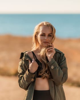 Portrait blonde sea cape. A calm young blonde in an unbuttoned khaki raincoat stands on the seashore, under the raincoat there is a black skirt and top