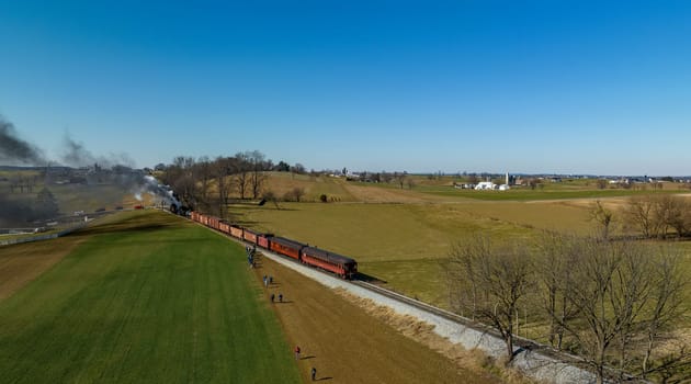 Ronks, Pennsylvania, February 18, 2023 - An Aerial View of a Steam Double-Header Freight, Passenger Train Combo. Picking Up Passengers in the Middle of Country Farmlands, on a Sunny Winter Day