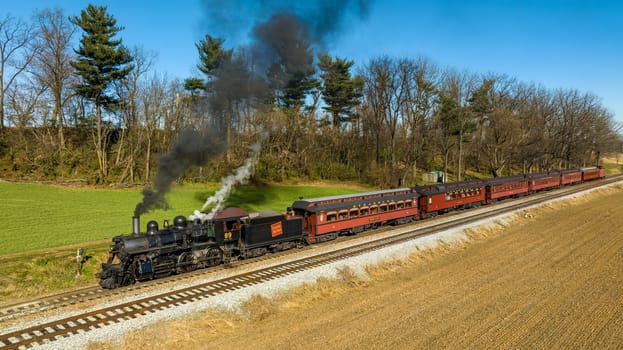 Ronks, Pennsylvania, November 26, 2022 - A Drone View of a Restored Steam Passenger Train Traveling Thru Farmlands Pulling Up to a Small Station on an Autumn Day
