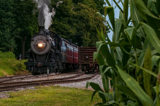 Front View of a Steam Locomotive Blowing Smoke and Steam With Freight and Corn Stalks on the Right