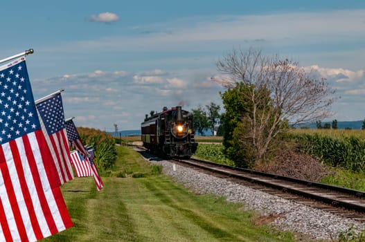 Restored Antique Steam Passenger Train Approaches a Fence With Gently Waving American Flags on It