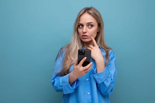 thoughtful carefree enchanted blonde woman with a smartphone in her hands on a blue bright background