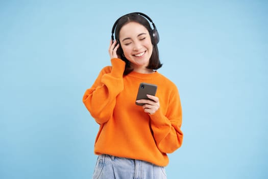 Happy young modern woman with headphones and smartphone, listening to music on mobile phone, enjoys favourite podcast, blue background
