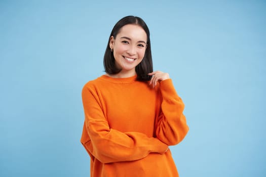Cute smiling asian woman, student in orange shirt, looks cute against blue background. Student and lifestyle concept