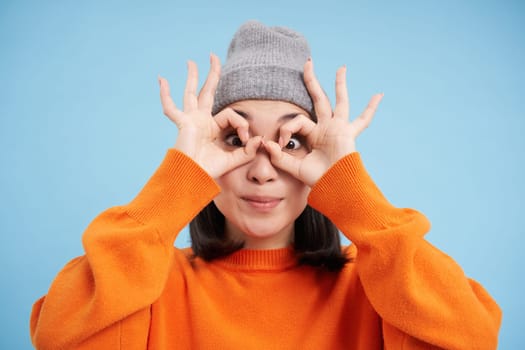 Close up portrait of funny Chinese girl, looks through hand glasses with surprised face expression, standing over blue background