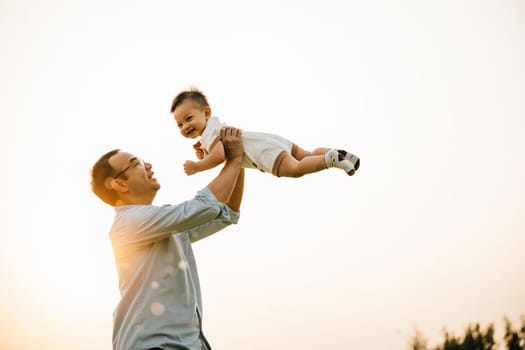 A happy dad and his toddler son share a playful moment of freedom and joy in the park, throwing him up in the air on a sunny summer day. Family love and happiness captured in a photograph