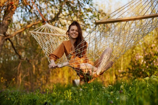 a joyful woman with long hair is lying in a hammock in an orange dress and happily smiling at the camera. A photo taken on the street on the theme of recreation in the country