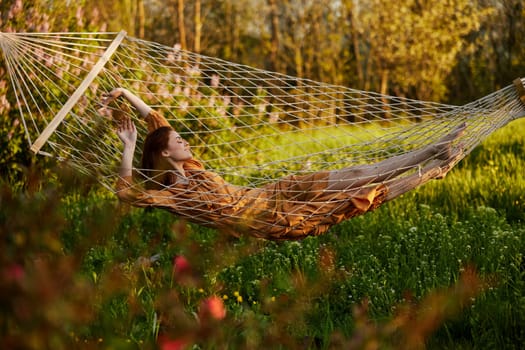 a beautiful woman is resting in nature lying in a mesh hammock in a long orange dress looking to the side, arms outstretched. Horizontal photo on the theme of recreation