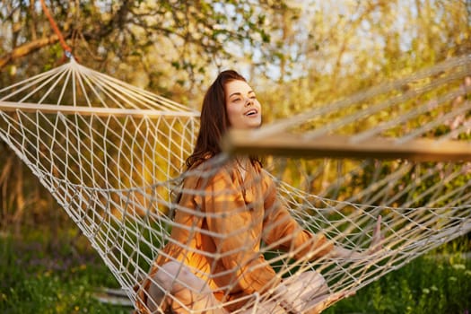 a joyful woman is sitting in a mesh hammock in nature relaxing and enjoying the rays of the setting sun on a warm summer day. Horizontal photo on the theme of outdoor recreation