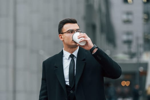 With cup of drink. Businessman in black suit and tie is outdoors in the city
