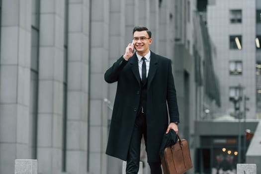 With smartphone. Businessman in black suit and tie is outdoors in the city