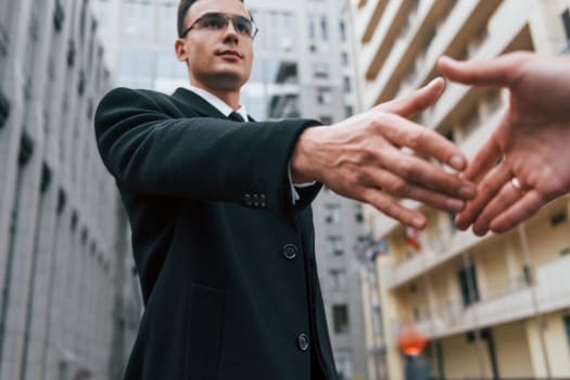 Doing handshake. Businessman in black suit and tie is outdoors in the city