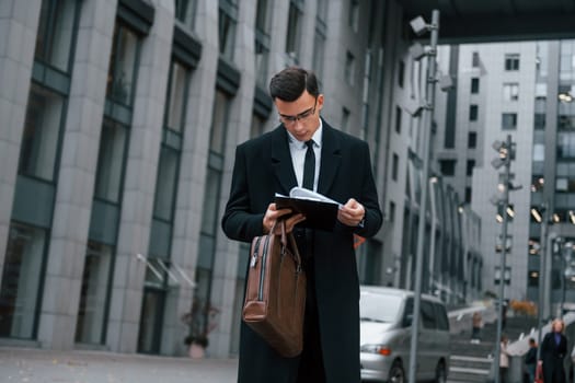 With notepad in hands. Businessman in black suit and tie is outdoors in the city