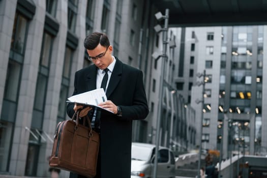 Holding documents. Businessman in black suit and tie is outdoors in the city