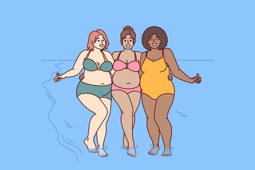 Cheerful plus size women dressed in swimsuit stand hugging on sunny beach near sea water