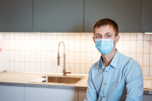 young guy sits in a shirt and a mask in the kitchen