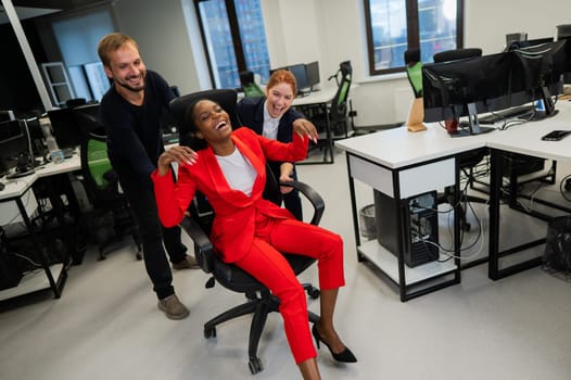 Caucasian red-haired woman, bearded caucasian man rolled African American young woman on office chair. Colleagues have fun at work.