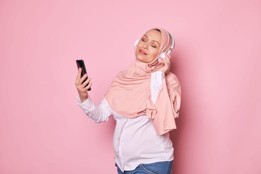 Delightful Muslim pregnant woman in pink hijab, enjoys soothing music on headphones, posing with eyes closed and a phone