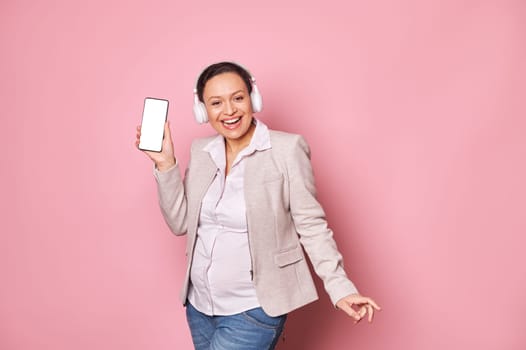 Smiling happy pregnant woman listens to music on headphones, dances over isolated pink background