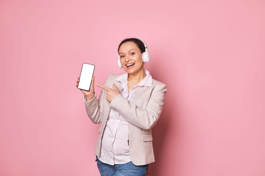 Pregnant woman in headphones, smiling at camera, showing smartphone with blank white screen with ad space for mobile app