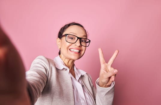 Happy woman smiles and shows peace sign, makes self-portrait on smartphone in her outstretched hands, isolated on pink