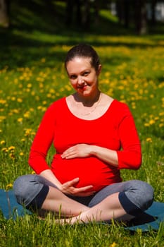 Pregnancy yoga exercise - pregnant woman doing asana Sukhasana easy yoga pose holding her abdomen outdoors on grass lawn with dandelions in summer