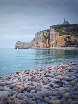 Sightseeing view to Etretat coastline with the famous Notre-Dame de la Garde chapel on the Amont cliff. Pebble beach washed by Atlantic ocean waters, Normandy, France