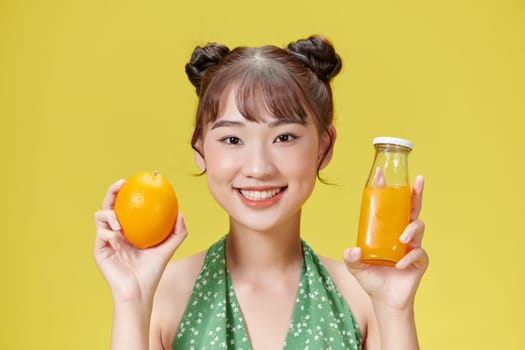 Young woman over isolated yellow background holding an orange and an orange juice