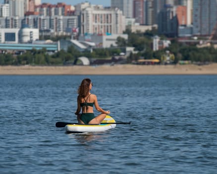 Caucasian woman is riding a SUP board on the river in the city. Summer sport.