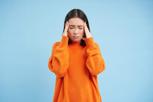 Frustrated asian woman holds hands on head, has headache, looks miserable, cant handle pressure, feels distressed, stands over blue background. Mental health concept
