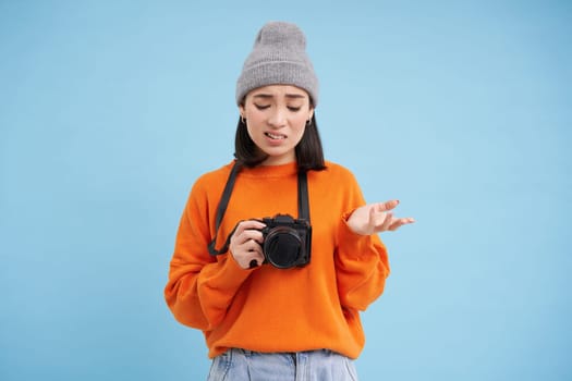 Portrait of asian woman in hat, holding digital camera with confused face, unprofessional photographer doesnt know how to take pictures on digicam, blue background