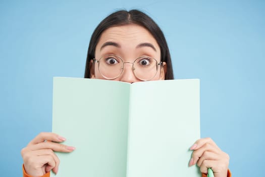 Cute asian woman in glasses, hides her face behind notebook and smiles with eyes, curious gaze, stands over blue background. Students and education concept