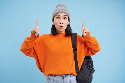Portrait of asian girl with shocked face, pointing fingers up and say wow, drops jaw, looks impressed, stands over blue background