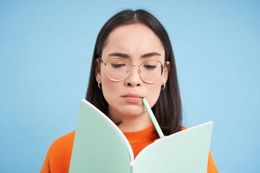 Portrait of thinking asian woman in glasses, solves task in notebook, writes down exercise, student learning, doing homework, blue background