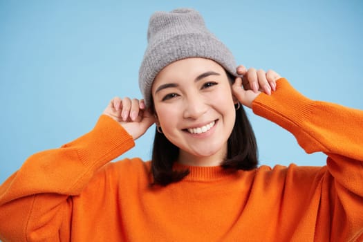 Close up portrait of smiling asian woman in warm hat, looking happy and cute at camera, has clear natural skin, stands over blue background