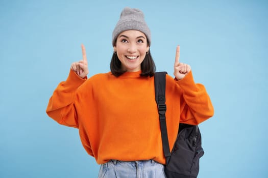 Smiling beautiful korean woman, pointing finger up, shows advertisement on top, wears street outfit and backpack, blue background