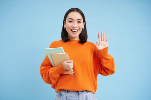 Friendly young woman, student says hello, stands with notebooks and waves hand to greet you, stands over blue background
